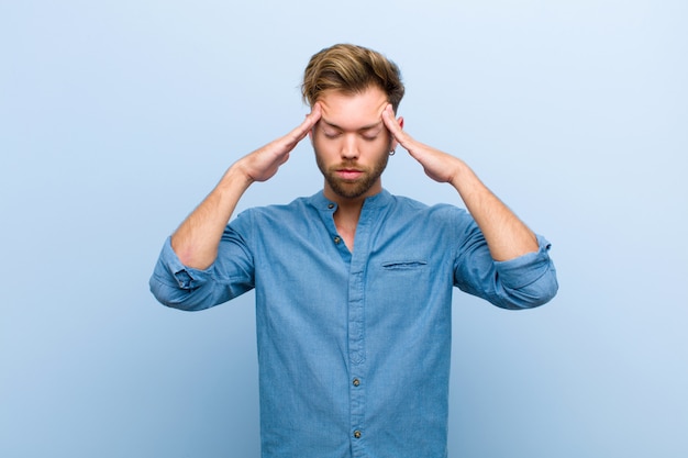 Young businessman looking concentrated, thoughtful and inspired, brainstorming and imagining with hands on forehead against blue wall