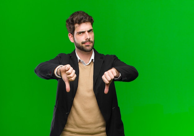 Young businessman looking sad, disappointed or angry, showing thumbs down in disagreement, feeling frustrated against green 