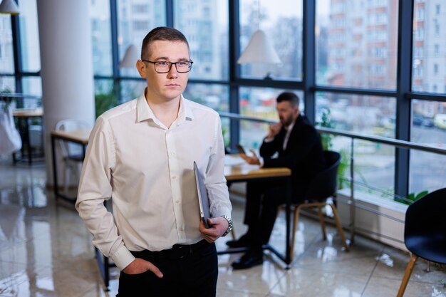 A young businessman man in glasses and a white shirt with a laptop in his hands stands in the office and works