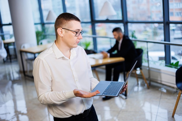 A young businessman man in glasses and a white shirt with a laptop in his hands stands in the office and works