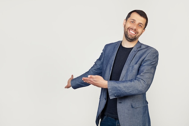 Young businessman man with a beard in a jacket, inviting to enter, smiling naturally with an open hand. Portrait of a man on a gray background.