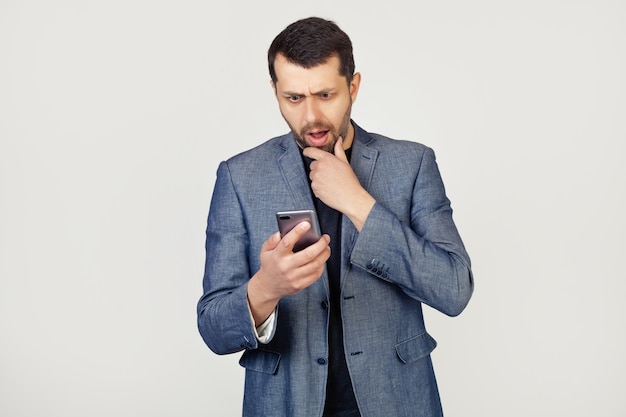 Young businessman man with a beard in a jacket using a smartphone scared shocked with a surprised face