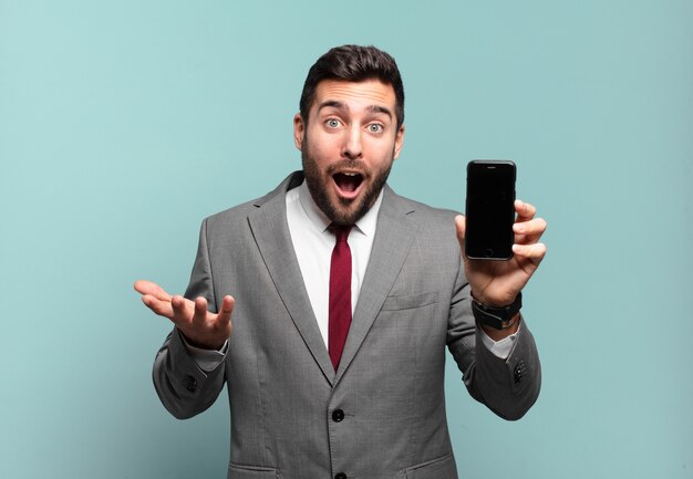 Молодой бизнесмен с открытым ртом изумлен, шокирован и изумлен невероятным сюрпризом и показывает экран своего телефона