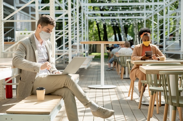 Молодой бизнесмен в защитной маске и формальной одежде смотрит на дисплей ноутбука, сидя на скамейке в летнем кафе в солнечный день
