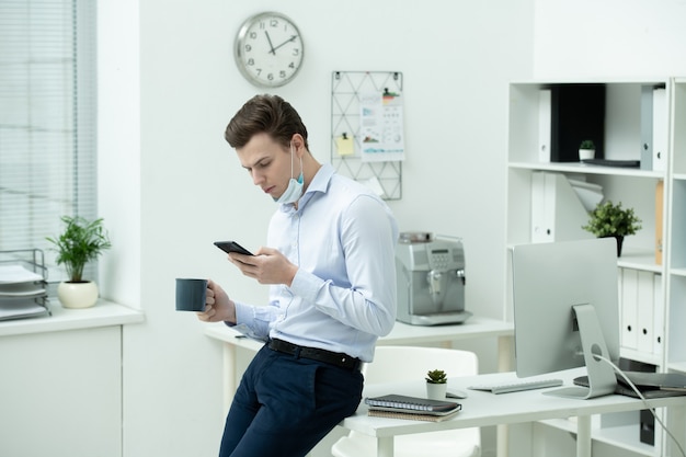 Young businessman in protective mask and formalwear using smartphone while sitting on desk with office supplies and having tea or coffee