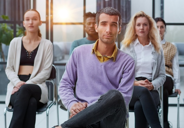 A young businessman in a purple sweater sits against the background of his colleagues in a modern of