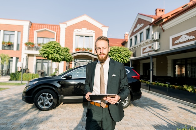 Молодой бизнесмен или менеджер по продажам в темном костюме стоял на открытом воздухе во дворе бизнес-центра с новым черным автомобилем