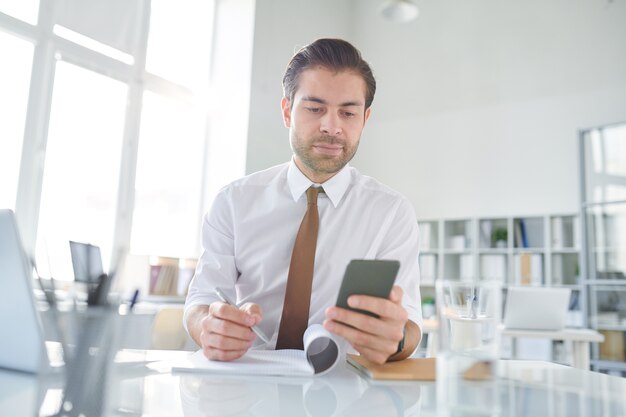 Young businessman scrolling in smartphone through business texts and writing down some new ideas