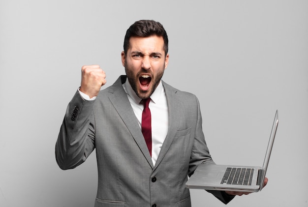 Young businessman shouting aggressively with an angry expression or with fists clenched celebrating success and holding a laptop