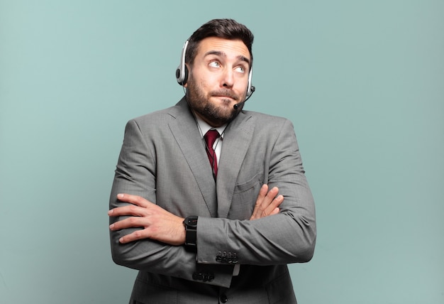 Young businessman shrugging, feeling confused and uncertain, doubting with arms crossed and puzzled look telemarketing concept