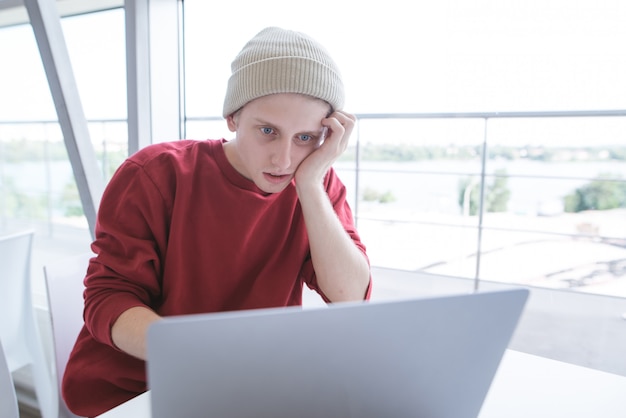 Молодой бизнесмен сидит у окна работает на ноутбуке и выглядит сосредоточенным на экране
