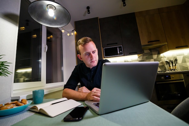 A young businessman sits at a table, holds a smartphone in his hand, uses a laptop and takes notes in his notebook. The guy is working, studying. Online marketing, education, e-learning, e-commerce.