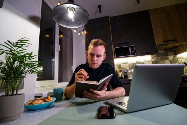 A young businessman sits at a table, holds a smartphone in his hand, uses a laptop and takes notes in his notebook. The guy is working, studying. Online marketing, education, e-learning, e-commerce.