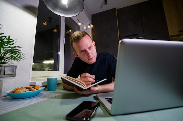 A young businessman sits at a table, holds a smartphone in his hand, uses a laptop and takes notes in his notebook. The guy is working, studying. Online marketing, education, e-learning, e-commerce.