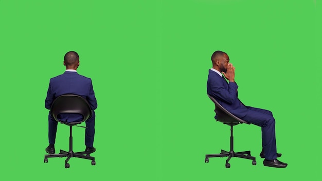 Young businessman sitting on chair and waiting, wearing formal office suit and posing over full body greenscreen backdrop. Male startup employee feeling optimistic in studio, stylish worker.