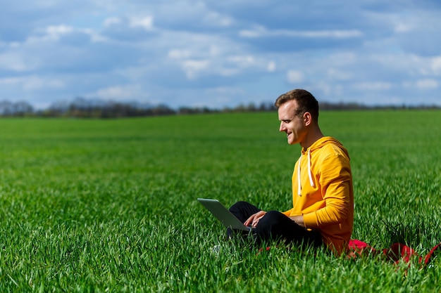 Young businessman sitting on green grass and using laptop computer. Handsome man working with computer in park at sunny summer day. Outdoors nature journey and relaxation. Freelance work concept