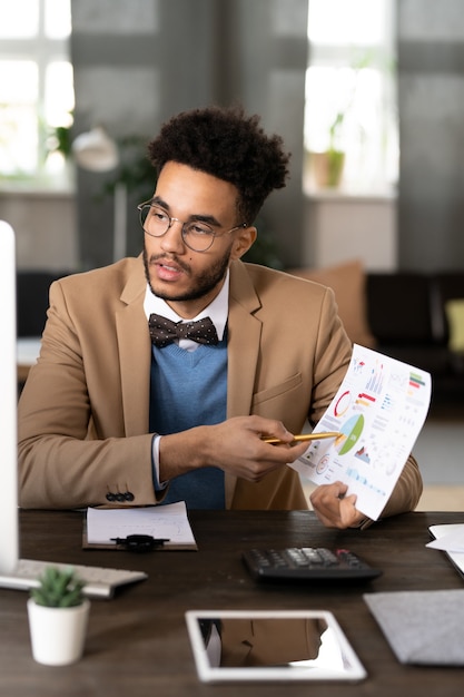 Photo young businessman sitting at his workplace and pointing at financial report in his hands during online presentation on computer