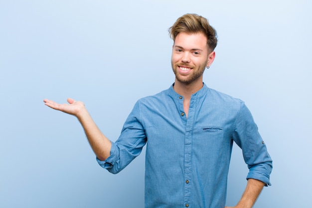 Young businessman smiling, feeling confident, successful and happy, showing concept or idea on copy space on the side against blue background