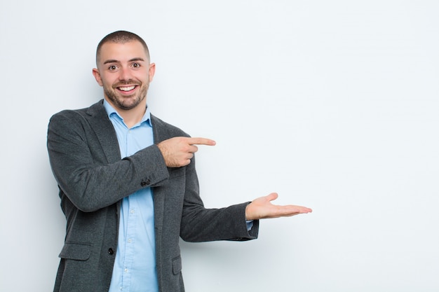 Young businessman smiling, feeling happy, carefree and satisfied, pointing to concept or idea on copy space on the side on flat wall
