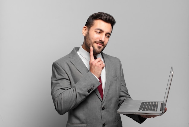 Young businessman smiling happily and daydreaming or doubting, looking to the side and holding a laptop