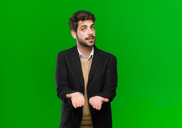 Young businessman smiling happily with friendly, confident, positive look, offering and showing an object or concept against green wall