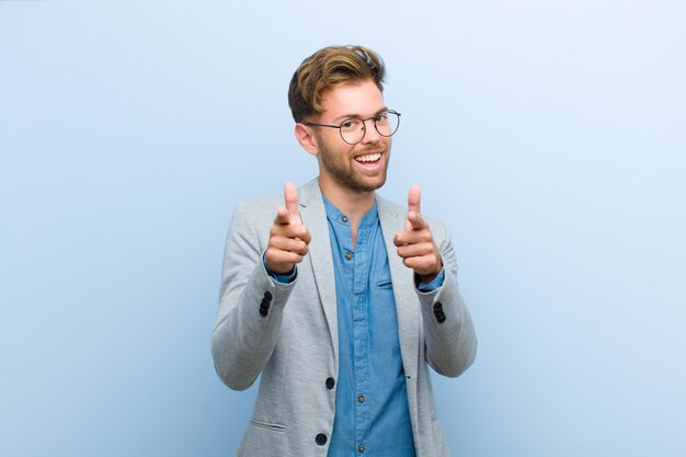 Photo young businessman smiling with a positive, successful, happy attitude pointing, making gun sign with hands against blue wall