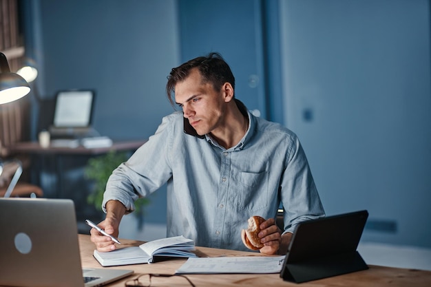 Young businessman snacking on a hamburger while working on a\
new project