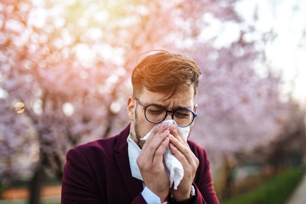 Young businessman sneezing in park. Allergy, flu, virus concept.