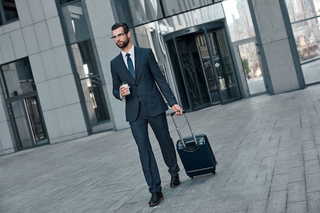 A young businessman spectacled crosses road with coffe and suitcase