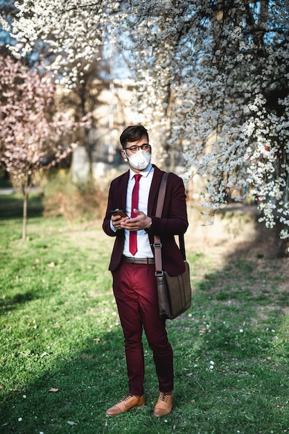 Young businessman standing in park, using phone and wearing protective mask. healthcare, virus protection, allergy protection concept