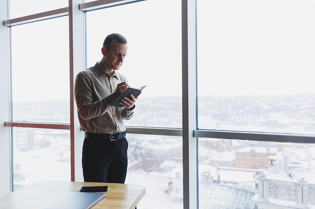 A young businessman stands at a panoramic window in a\
skyscraper and makes notes in his notebook with a pen a man in a\
shirt and trousers of european appearance