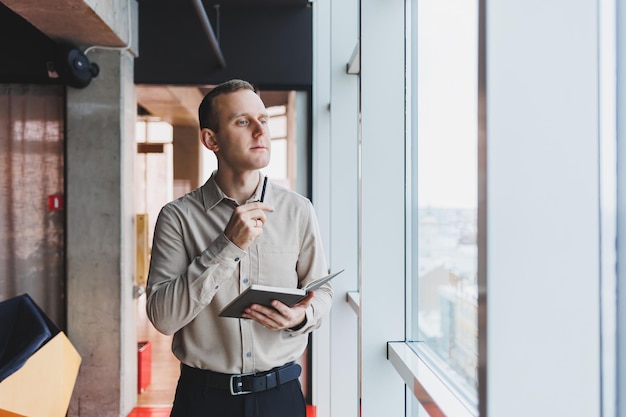 A young businessman stands at a panoramic window in a skyscraper and makes notes in his notebook with a pen A man in a shirt and trousers of European appearance