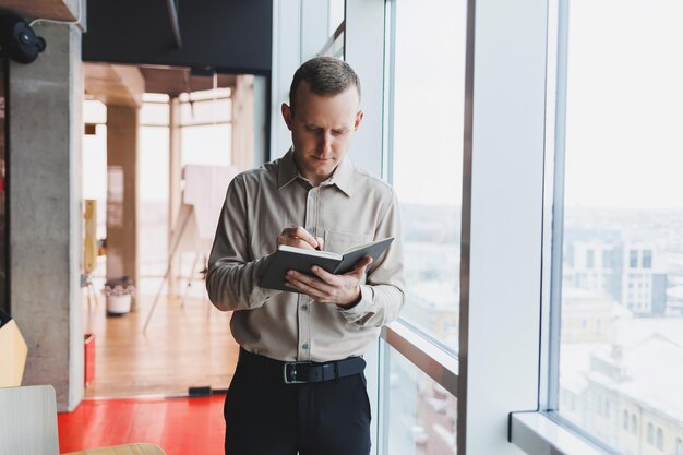 A young businessman stands at a panoramic window in a skyscraper and makes notes in his notebook with a pen A man in a shirt and trousers of European appearance