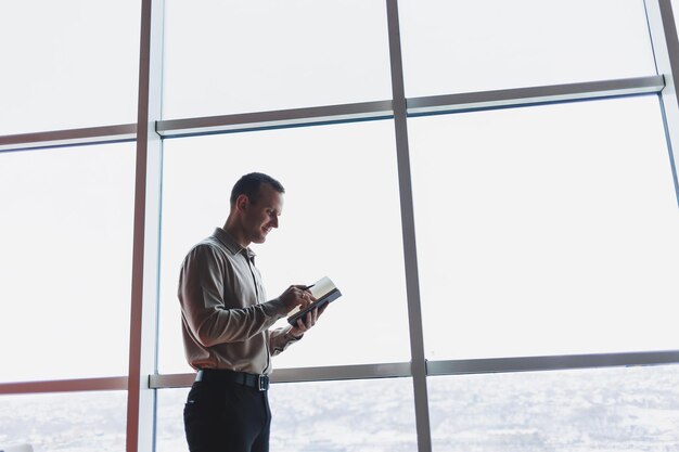 Un giovane uomo d'affari si trova davanti a una finestra panoramica in un grattacielo e prende appunti sul suo taccuino con una penna un uomo in camicia e pantaloni di aspetto europeo