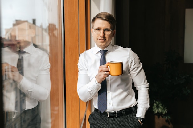 Il giovane uomo d'affari sta con una tazza di caffè vicino alla finestra del suo ufficio