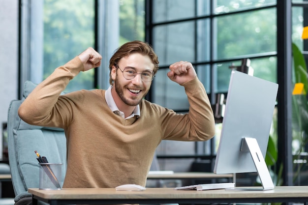 A young businessman student freelancer programmer sits in the office at the table and celebrates