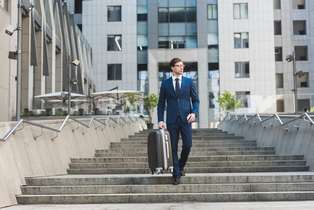 Young businessman in stylish suit with luggage and flight tickets going down stairs in business