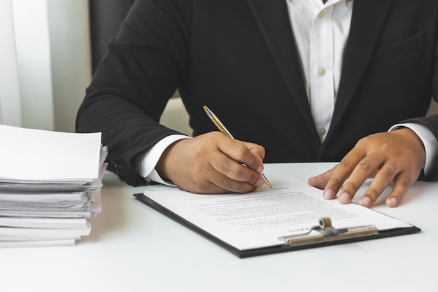 A young businessman in a suit is signing documents for a\
variety of tasks in the office. many paperwork that is not\
finished. documents in the company about finance and information of\
the company.