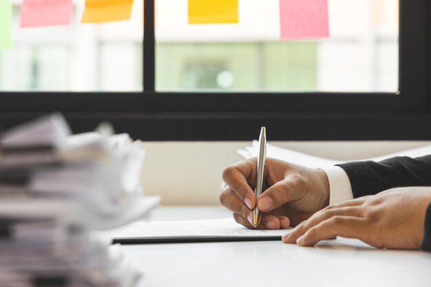 A young businessman in a suit is signing documents for a\
variety of tasks in the office. many paperwork that is not\
finished. documents in the company about finance and information of\
the company.