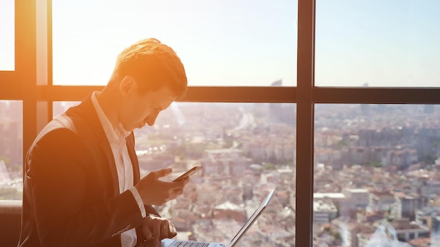 Young businessman in suit is working with phone and laptop in office. He is sitting near the window with panoramic city view, copy space