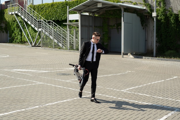 Young businessman in a suit looks at his watch and carries an electric scooter in his hands