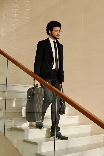 Young businessman in suit moving down the stairs with suitcases in office building