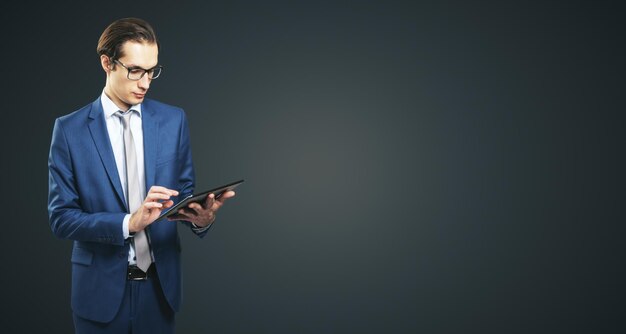 Young businessman in suit with digital tablet on dark grey background with place for your text mock up