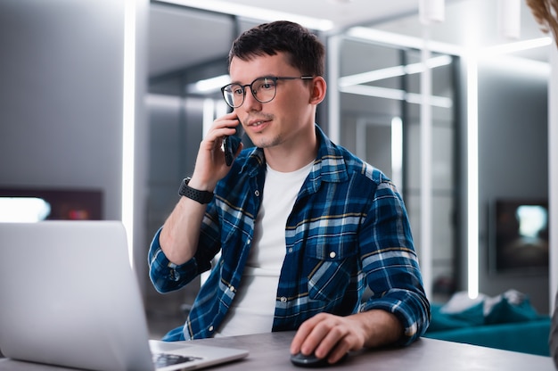 Young businessman talking on phone sitting at kitchen table, busy entrepreneur working distantly from home with laptop papers speaking on mobile.