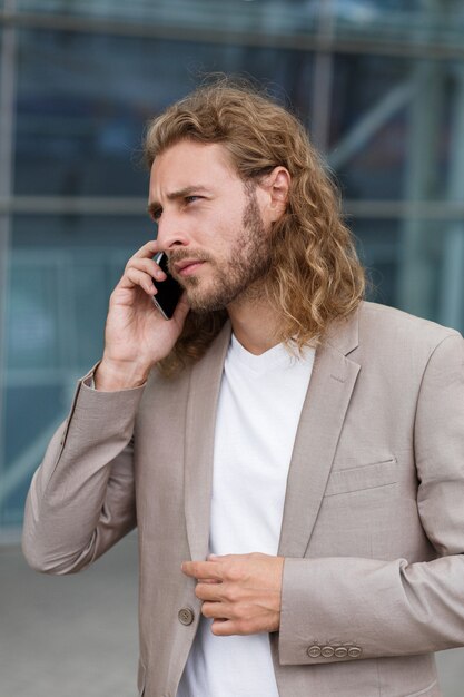 Young businessman talking on the phone, standing on city street near business center. Successful manager makes a call. Portrait of handsome curly smiling man in casual wear holding smartphone.