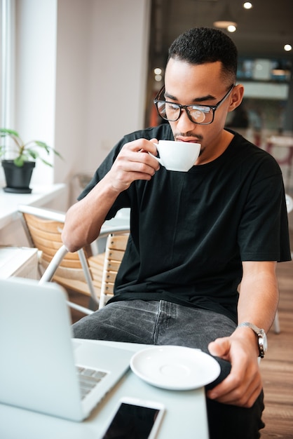 young businessman wearing glasses sitting near window and typing by laptop computer while drinking coffee