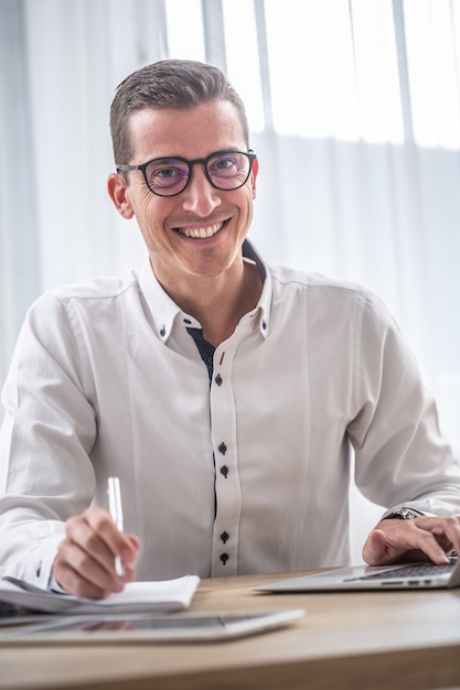 Foto il giovane uomo d'affari con gli occhiali sorride alla telecamera mentre è seduto alla scrivania dell'ufficio.