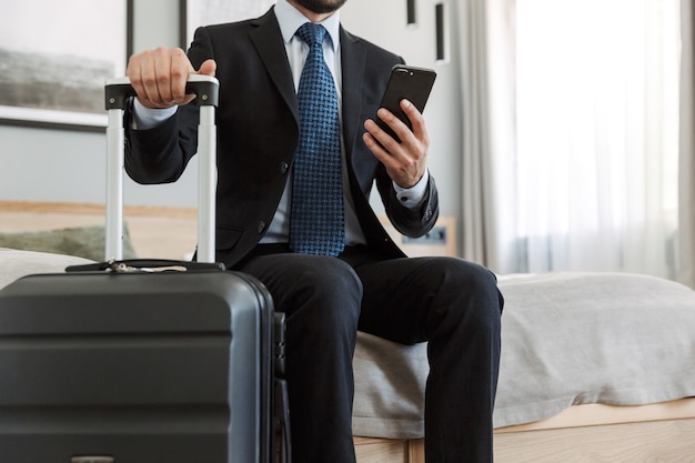 young businessman wearing suit sitting at the hotel room, using mobile phone while carrying suitcase