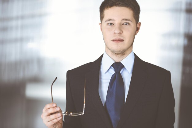 Young businessman wearing suit, with glasses, while standing in the office. Business success concept nowadays.