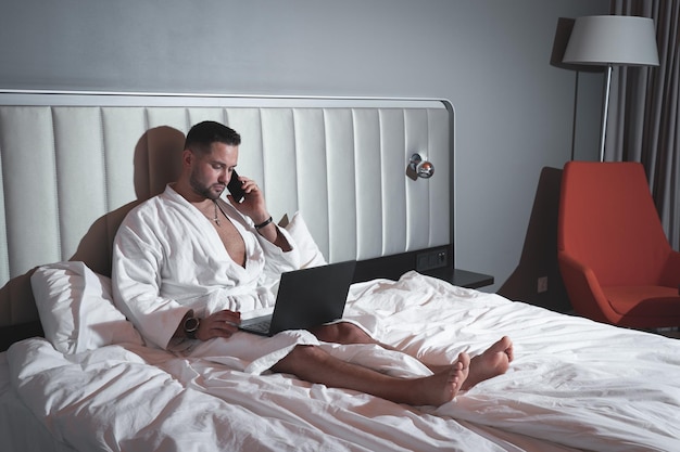 A young businessman in a white coat sitting on a bed talking on a mobile phone and working